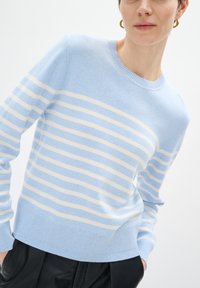 Maglione in lana azzurro chiaro con strisce bianche orizzontali, scollatura rotonda, polsini e orlo a costine, texture liscia. Il modello è posizionato leggermente di lato.