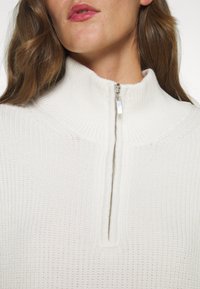 Femme portant un pull en maille côtelée blanche avec un col à demi-fermeture éclair argentée, montrant ses lèvres et ses cheveux bruns ondulés.