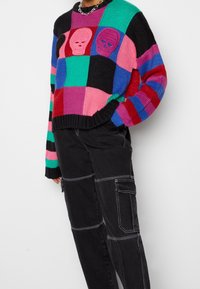Maglione lavorato a maglia colorato con motivi di teschi in un motivo a scacchi rosa, verde e nero; abbinato a pantaloni cargo neri con cuciture dettagliate.