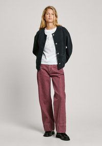 Schwarze Cordjacke mit silbernen Knöpfen, weißes Grafik-T-Shirt und burgunderfarbene, weit geschnittene Jeans mit kontrastierenden Nähten, kombiniert mit schwarzen Schuhen.