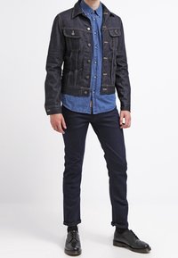 Tumma farkkutakki, jossa on rakennettu muotoilu, rintataskut, hopeiset napit ja erottuva ommel, kerroksittain vaaleansinisen paidan päällä.