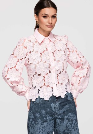 Donna che indossa una camicetta in pizzo floreale rosa chiaro a maniche lunghe con colletto e bottoni sul davanti, abbinata a jeans scuri con motivo floreale.