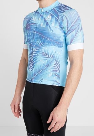 Hombre que lleva un maillot de ciclismo azul con patrones de hojas de palma y pantalones cortos de ciclismo negros, de pie con los brazos relajados a los lados.