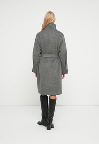 Grauer Wollmantel mit hohem Kragen, tailliertem Bund, langen Ärmeln und knielangem Design. Glatte Textur und dezent meliertes Muster.