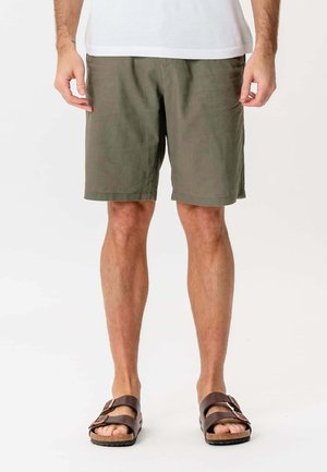 Mand iført olivengrønne shorts, hvid skjorte og brune sandaler, stående med afslappede hænder ned langs siden på hvid baggrund.