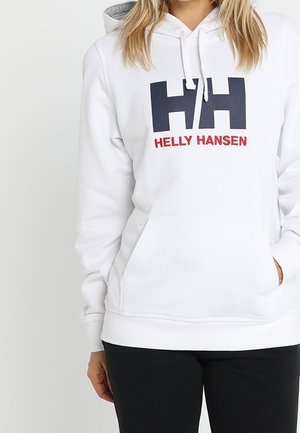 Mujer con una sudadera con capucha blanca de Helly Hansen con un gran logo negro y rojo, combinada con pantalones negros, de pie frente a un fondo liso.