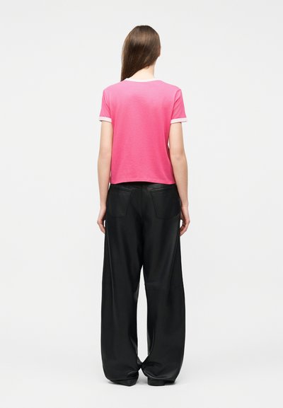 Γυναίκα με μακριά καστανά μαλλιά που φοράει ροζ κοντομάνικη μπλούζα και μαύρο παντελόνι φαρδιάς γραμμής, στέκεται με την πλάτη στην κάμερα.