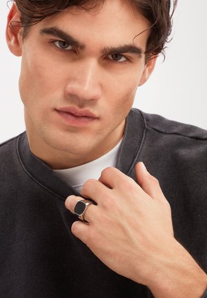 Młody mężczyzna o ciemnych włosach nosi czarną bluzę i srebrny pierścień z czarnym kwadratowym kamieniem na prawej ręce w pobliżu szyi.