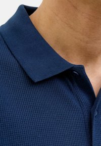 Granatowa koszulka polo z textured, waflowym splotem. Posiada klasyczny kołnierzyk i zapięcie na guziki, zbliżenie na dekolt.