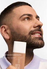 Bottiglia di fondotinta marrone con cappuccio bianco etichettata "Fondotinta Ogni Singolo Giorno SPF 50". Pelle con leggera barba, indossa una polo viola chiaro.