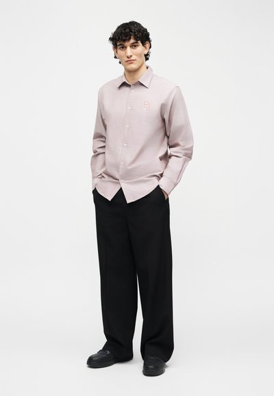 Νεαρός με σγουρά μαλλιά, ντυμένος με ανοιχτό ροζ πουκάμισο με κουμπιά και λογότυπο "EA" και φαρδιά μαύρα παντελόνια, στέκεται με τα χέρια στις τσέπες.