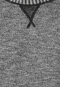 Tissu de pull en tricot gris présentant un motif texturé. Des coutures noires forment un 'X' au niveau de l'encolure, avec un design de col côtelé.
