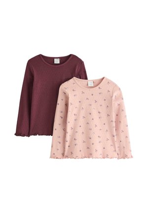 Ung pige med langt hår i lyserød blomstret langærmet skjorte, løstsiddende mørkeblå jeans og røde sko, der står med hænderne i lommerne.