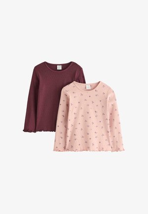 Ung pige med langt hår i lyserød blomstret langærmet skjorte, løstsiddende mørkeblå jeans og røde sko, der står med hænderne i lommerne.