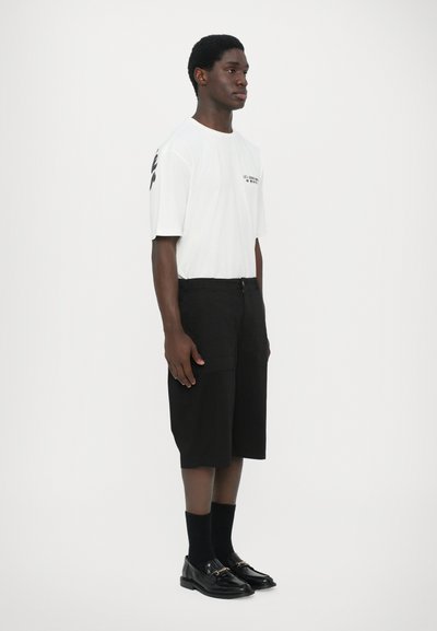 T-shirt blanc à manches courtes avec du texte noir, associé à un short noir jusqu'aux genoux et des mocassins noirs brillants avec des accents dorés.