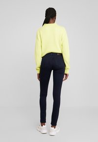 Mujer con cabello trenzado vistiendo un suéter amarillo claro, jeans skinny oscuros y zapatillas blancas, de espaldas frente a un fondo gris liso.