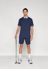 Πολό μπλουζάκι ναυτικού χρώματος με αντιθετικό γιακά, συνδυασμένο με κοντόπαντελόνι ναυτικού χρώματος. Αξεσουάρ περιλαμβάνουν μπαστούνι του γκολφ. Το μοντέλο φοράει λευκές κάλτσες και παπούτσια.