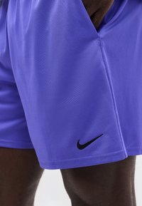 Κοντινό πλάνο ενός ατόμου που φοράει μοβ αθλητικά σορτς Nike με το χέρι στην τσέπη, παρουσιάζοντας υφή υφάσματος και λογότυπο κοντά στο τελείωμα.