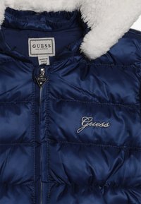 Granatowa kurtka puchowa z białym kołnierzem z polaru, zamek błyskawiczny z przodu oraz haftowanym logo "Guess" na piersi i nadrukiem na metce.