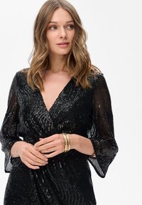 Kobieta z falującymi, jasnobrązowymi włosami, ubrana w czarną sukienkę z cekinami w serek i złote bransoletki, patrząca lekko w lewo na białym tle.