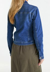 Sininen farkkutakki, jossa istuva malli, sivusaumat ja nappidetaljit; yhdistetty oliivinvihreään hameeseen, jossa on sivutaskut.