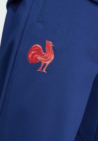 Blå atletiska byxor med en röd broderad tupp-logo. Tyget verkar slätt med en enhetlig struktur och en avslappnad passform.