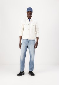 Jachetă din denim alb cu două buzunare pe piept, tricou bleu deschis, blugi bleu deschis și adidași negri. Căciulă albastră cu logo pe față.