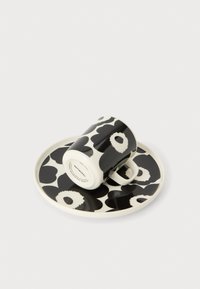 Κεραμικό φλιτζάνι με μοτίβο ασπρόμαυρα λουλούδια που ακουμπάει πλαγίως σε ένα ταιριαστό στρογγυλό πιάτο με απλό φόντο.