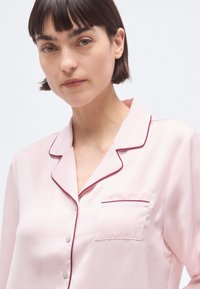 Haut de pyjama en soie rose avec col à revers, passepoil bordeaux, fermeture à boutons et une poche poitrine unique. Texture lisse, design élégant.