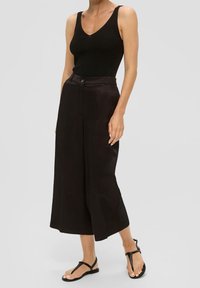 Černý žebrovaný top s hlubokým výstřihem ve tvaru V, v kombinaci s širokými vysokými černými kalhotami a plochými černými sandály. Hladká textura látky.