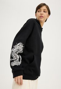 Noor inimene, kes kannab musta kapuutsiga pusa valge draakoni kujundusega varrukal, vaatab üle õla, seisab ühetaolise heleda tausta ees.