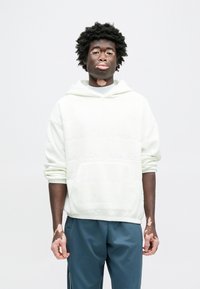 Sweat à capuche crème clair avec un motif texturé et une poche avant, associé à un pantalon de survêtement bleu foncé. Le tissu semble doux et décontracté.