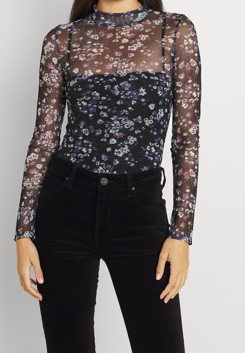 Langärmliger schwarzer Floral-Body mit transparentem Mesh-Stoff. Merkmale sind lila und weiße Blumen, Stehkragen und enganliegendes Design. Kombiniert mit schwarzen Hosen.