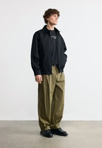 Νεαρός άνδρας που στέκεται, φοράει μαύρο μπουφάν πάνω από μαύρο t-shirt Calvin Klein, χαλαρό χακί παντελόνι και μαύρα παπούτσια, μπροστά σε απλό λευκό φόντο.