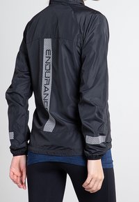 Chaqueta ligera negra con acentos reflectantes en la parte trasera y las mangas, hecha de un material suave y resistente al agua.