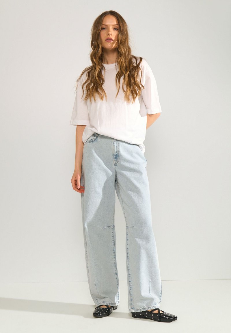 Femme aux longs cheveux ondulés portant un t-shirt blanc ample, un jean large bleu clair et des ballerines noires pointues avec des brides à la cheville.