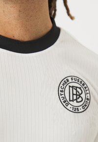 Fehér mez függőleges csíkokkal, fekete bordázott gallérral és kerek emblémával, amelyen a "Deutscher Fussball-Bund" és a 125-ös szám szerepel.