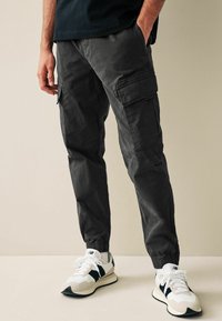 Dark gray cargo pants with elastic cuffs, featuring multiple side pockets and a relaxed fit. Worn with white and navy sneakers.