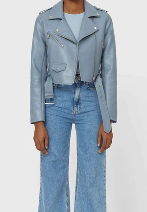 Personne portant une veste en cuir courte bleu clair avec des fermetures éclair argentées et une ceinture, associée à un jean large taille haute bleu.