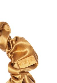 Scrunchie in raso dorato con una superficie liscia, dotato di un'etichetta in tessuto marchiata di colore coordinato, che mette in risalto un design arricciato per elasticità.