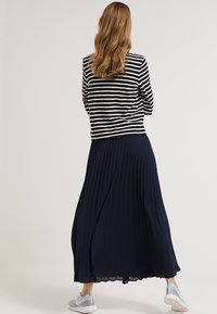 Gestreept top met lange mouwen in marineblauw en wit, gecombineerd met een vloeiende marineblauwe geplooide maxi-rok. Het model draagt lichtgekleurde sneakers.