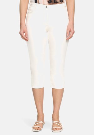 Kvinde iført tætsiddende hvide korte bukser og hvide flade sandaler med stropper, stående med arme afslappet langs siderne.