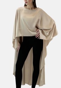 Beige, flödande kapp-topp gjord av ett mjukt tyg, med vida ärmar och asymmetrisk fåll, kombinerad med svarta leggings.