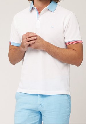 Biała koszulka polo z jasnoniebieskim kołnierzykiem i czerwonymi, paskowanymi akcentami na rękawach. Na piersi znajduje się małe logo. Noszona z jasnoniebieskimi spodniami.