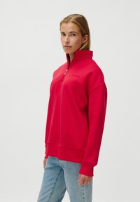 Pulover roșu din fleece cu fermoar pe jumătate, guler înalt și manșete riba. Prezintă un logo subtil pe pieptul stâng. Asortat cu blugi de culoare albastru deschis.