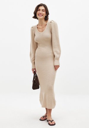 Abito lavorato a maglia beige con maniche lunghe a sbuffo, corpo aderente e orlo svasato. Una borsa marrone e infradito neri completano il look.
