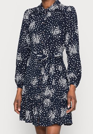 Femme portant une robe bleu marine avec des motifs de pois blancs dispersés, manches longues, boutonnée à l'avant, taille nouée et ourlet à volants.