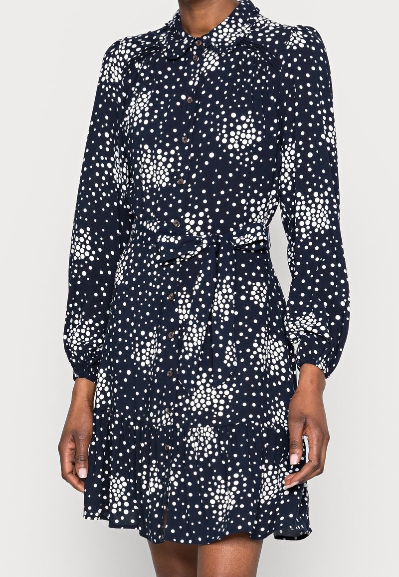 Femme portant une robe bleu marine avec des motifs de pois blancs dispersés, manches longues, boutonnée à l'avant, taille nouée et ourlet à volants.