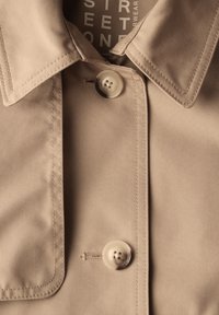 Beige trench coat -kangaskangas, jossa on sileä pinta, terävä kaulus ja kaksi läpinäkyvää nappia etuplaatassa.