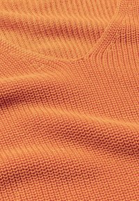 Dzianina w intensywnym pomarańczowym odcieniu, z teksturowanym wzorem, widocznymi ściegami i subtelnymi falistymi zagnieceniami.